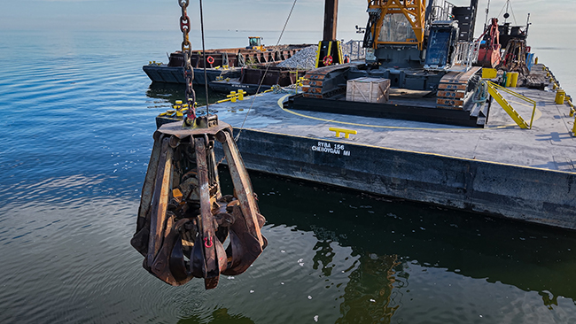 A close view of a grapple on the end of a crane, suspended just about the surface of the water. The grapple is about 15 feet tall and comprised of six arms that are folded in. In the background is a barge with a crane.